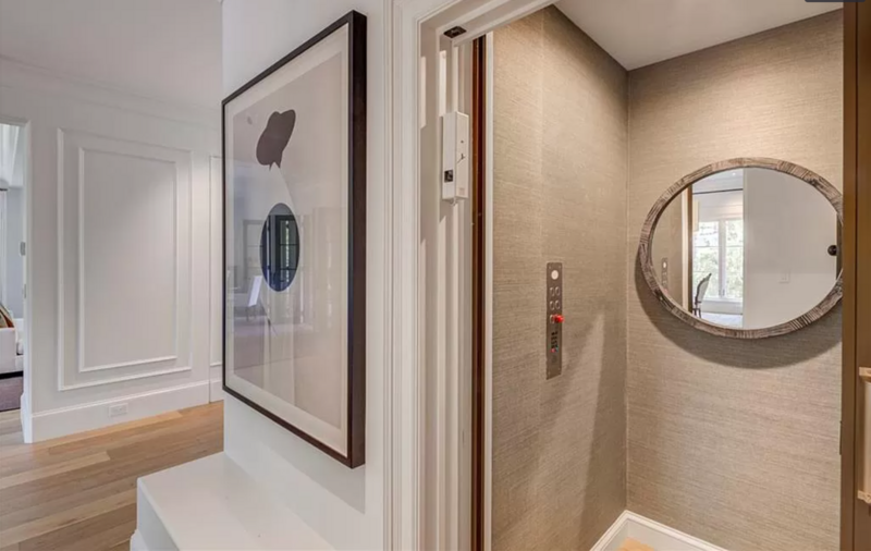 Hallway with elevator. Framed art on wall, circular mirror, beige walls, wood floor, white trim.