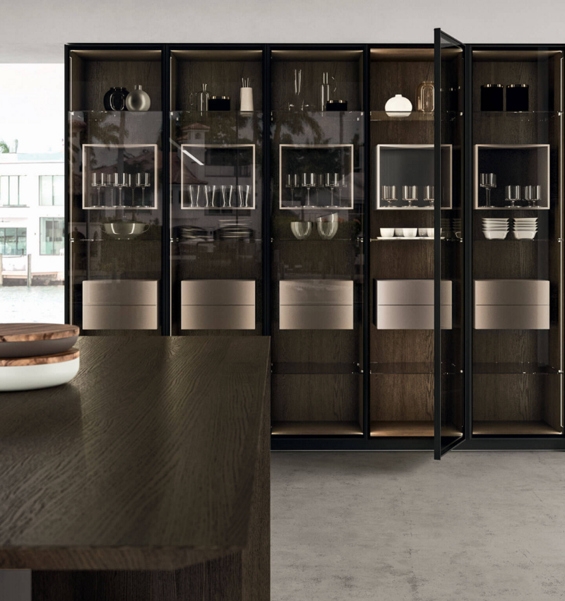 Modern kitchen cabinet with glass doors, displaying dishware. Dark wood and metallic accents; neutral tones.