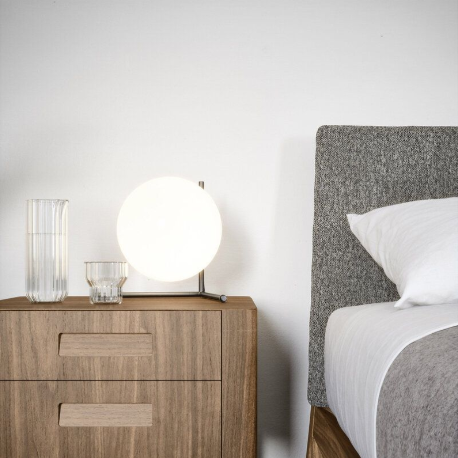 Nightstand with lamp, glasses, and a bed with a gray headboard and white bedding.
