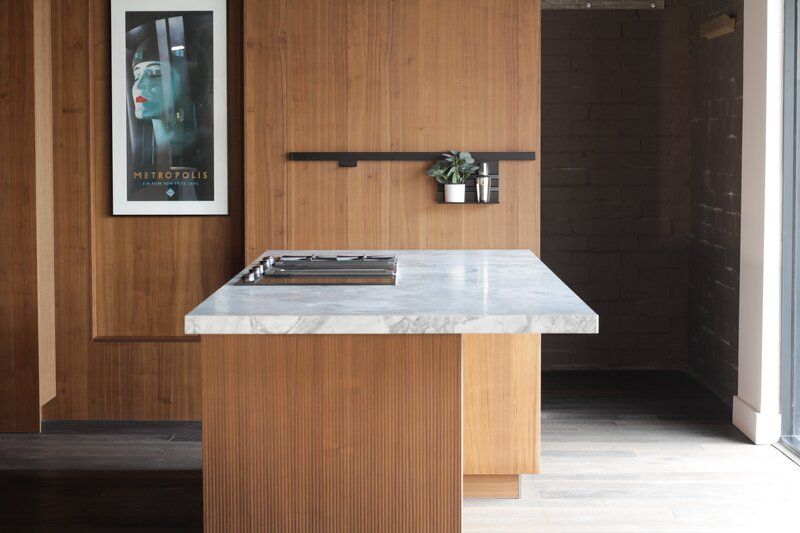 Modern kitchen with wood paneling, marble countertop island, built-in cooktop, and small shelf with decor.