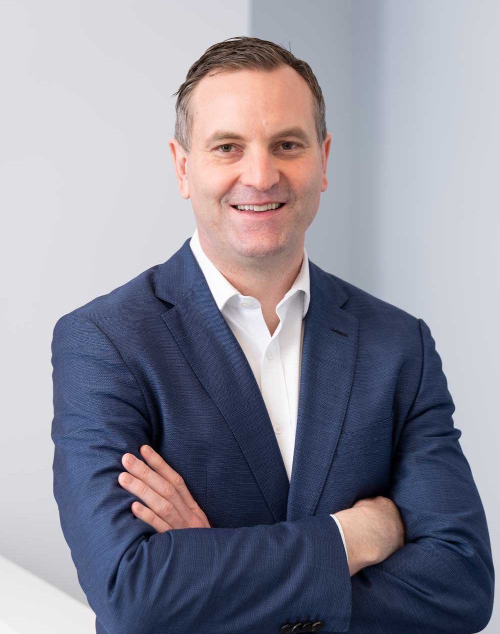 A man in a blue suit and white shirt is standing with his arms crossed and smiling.