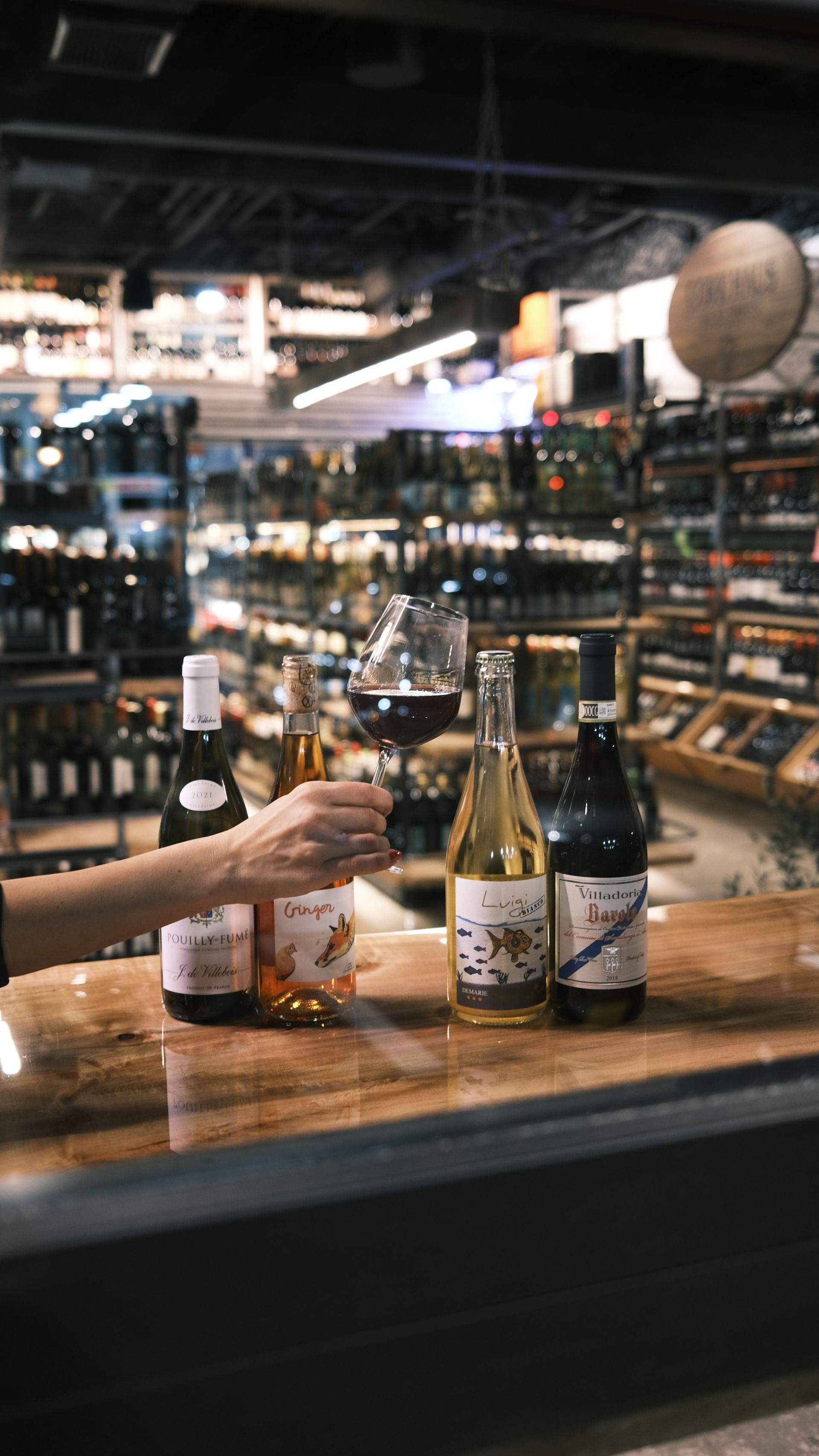 A person is holding a glass of wine while sitting at a table with bottles of wine.