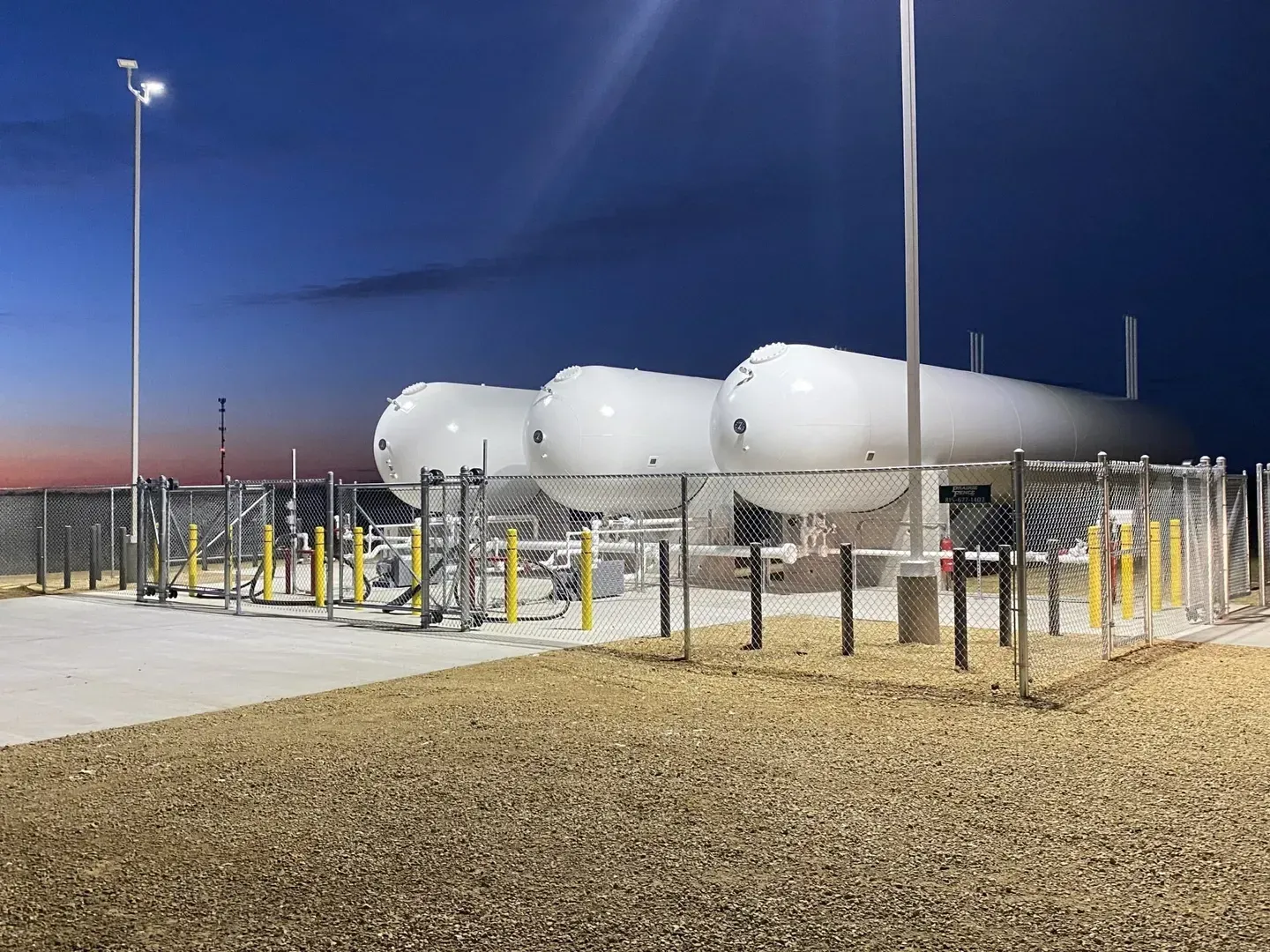 Large white industrial tanks behind a fence at dusk, lit by tall streetlights in a gravel lot