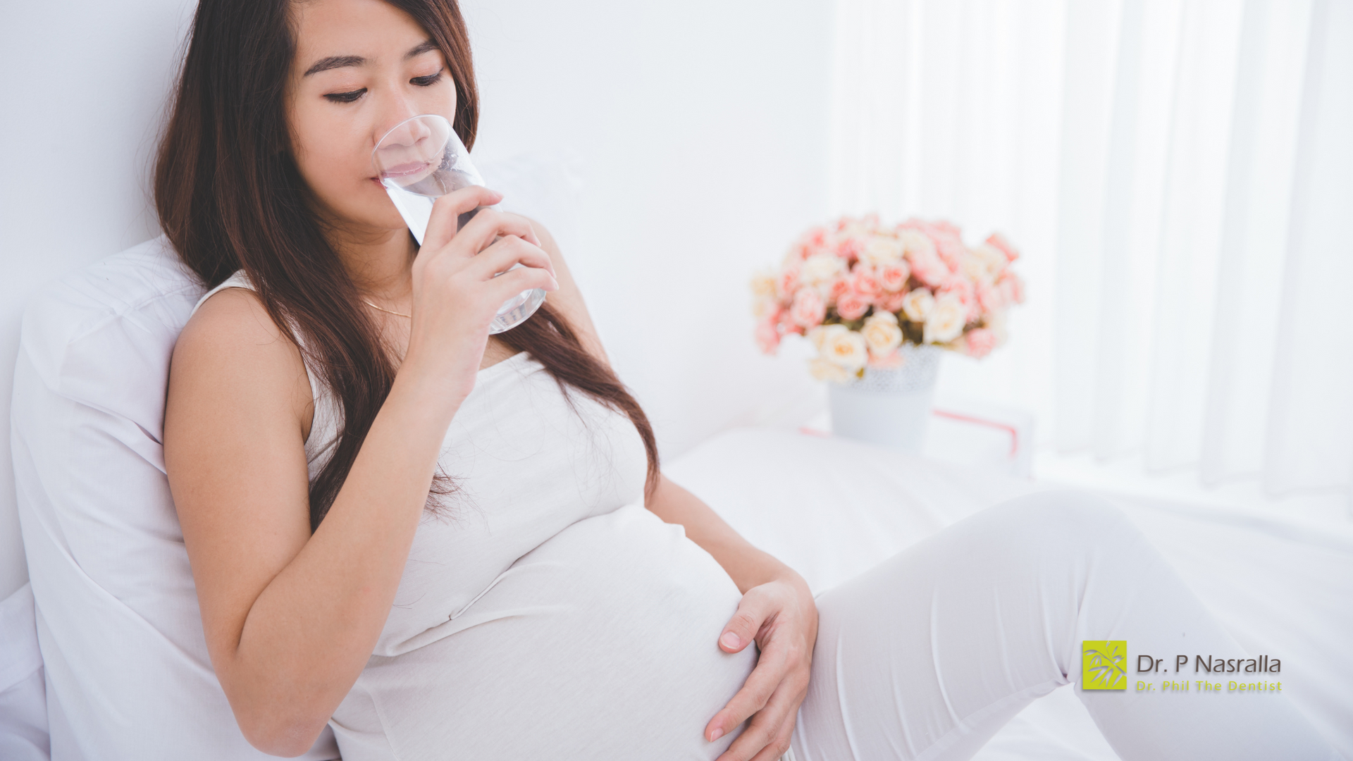 A pregnant woman is drinking water while sitting on a bed.