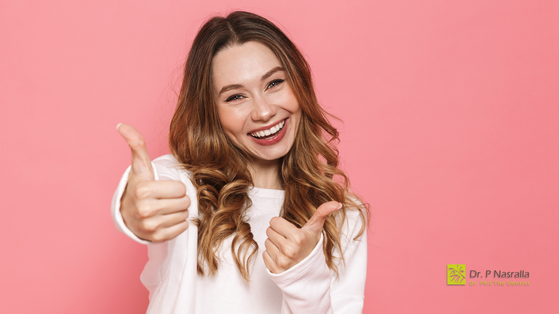Woman with long brown hair, giving double thumbs up, smiling against pink background.