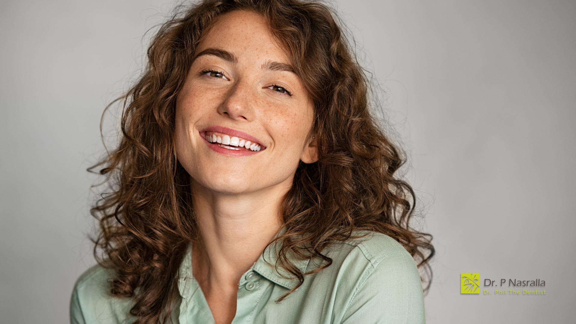 Woman with curly brown hair smiles, wearing a green shirt, against a gray backdrop.