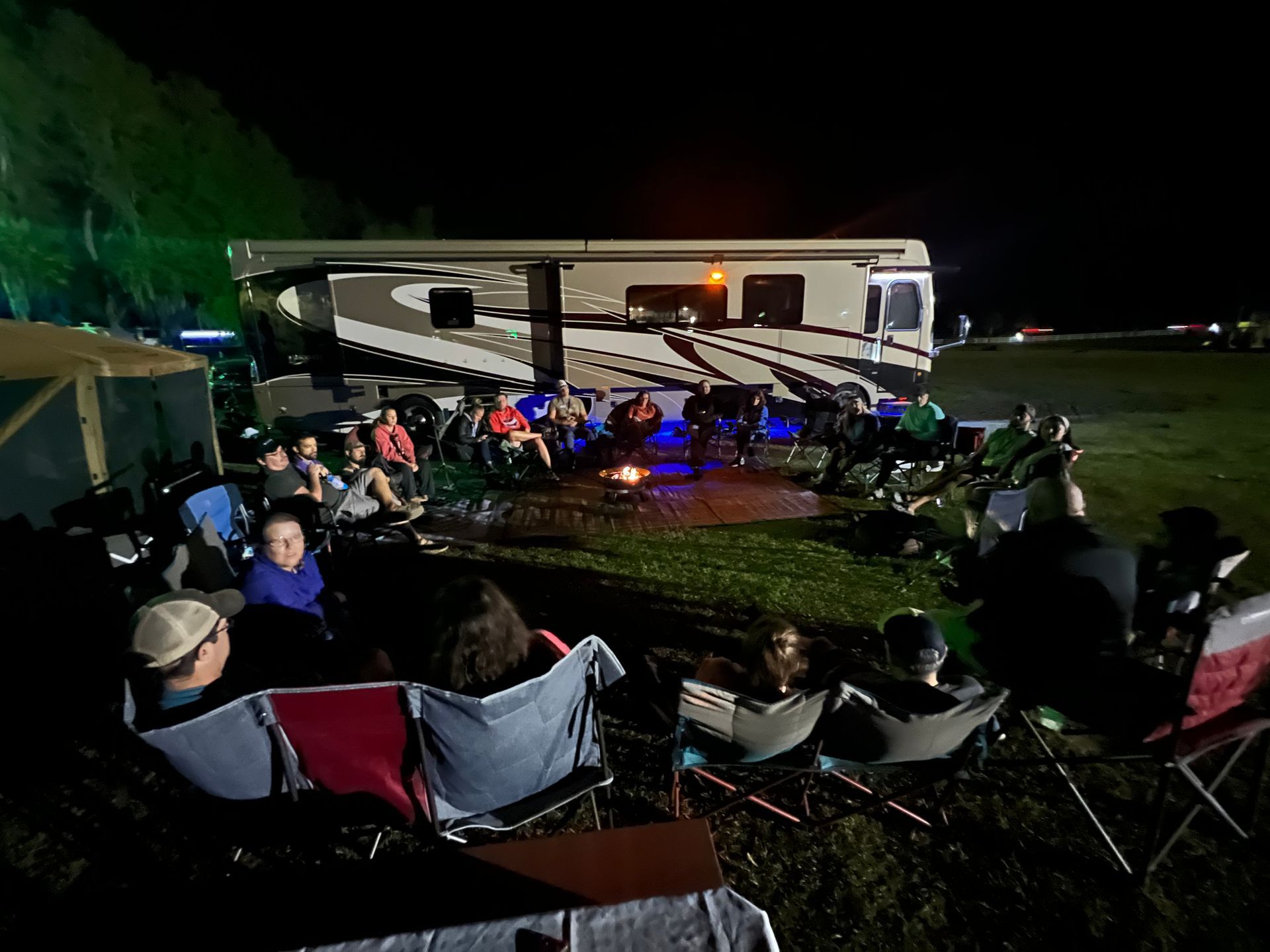 A group of people are sitting around a fire pit in front of a rv at night.