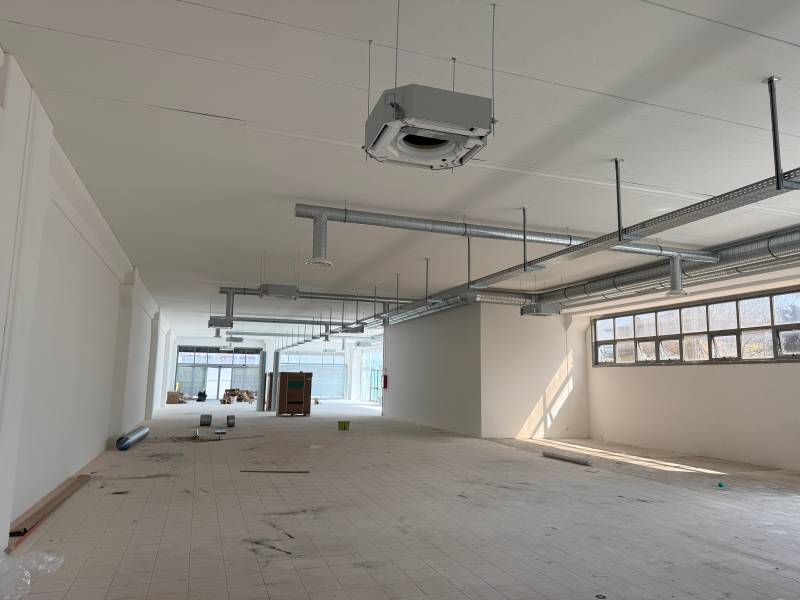 Stanza vuota in costruzione con pareti bianche, infissi a vista sul soffitto e grandi finestre.