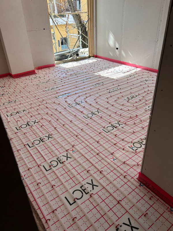 Stanza con impianto di riscaldamento a pavimento installato, con griglia e tubature bianche e rosse, vicino a una finestra.