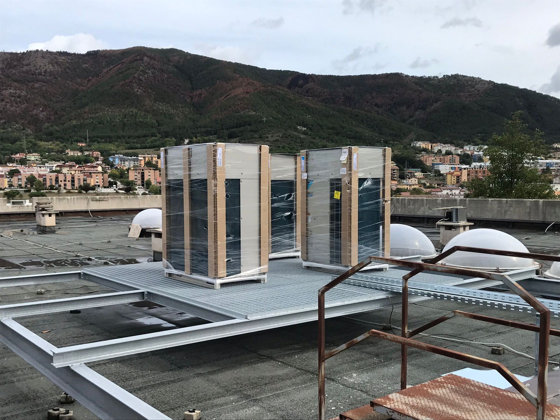 Tre unità HVAC preassemblate su una piattaforma metallica su un tetto, con una città e una montagna sullo sfondo.