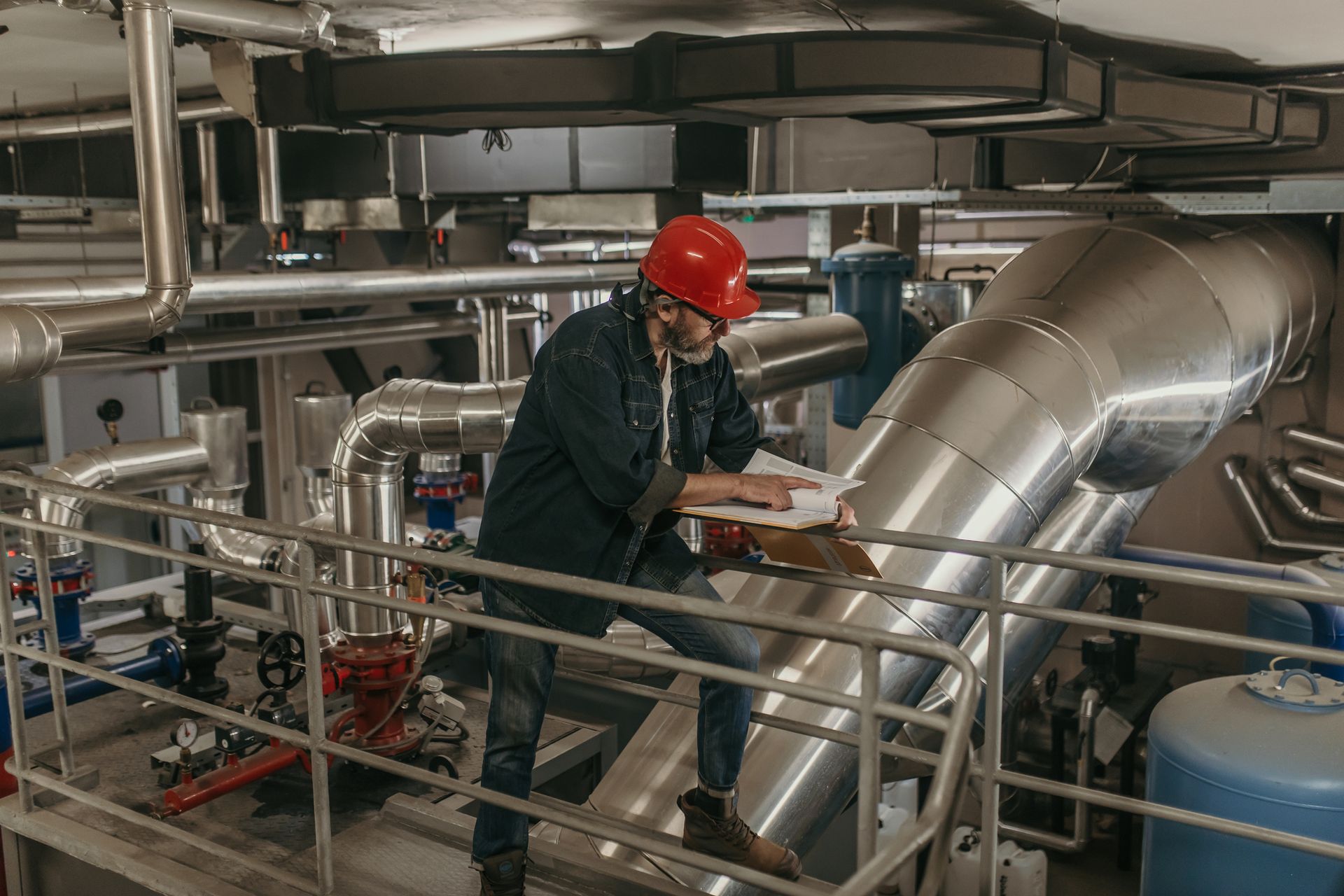 Ingegnere con casco rosso che ispeziona tubazioni e attrezzature industriali, scrivendo su un blocco appunti.