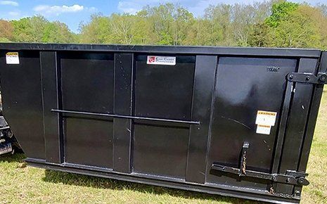 Black dumpster on a grassy field with trees in the background.