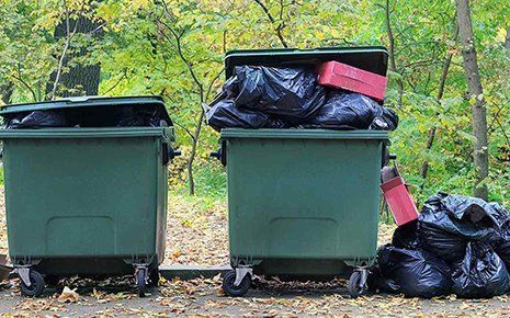 Two green dumpsters overflowing with black trash bags and a red box, in an outdoor setting.