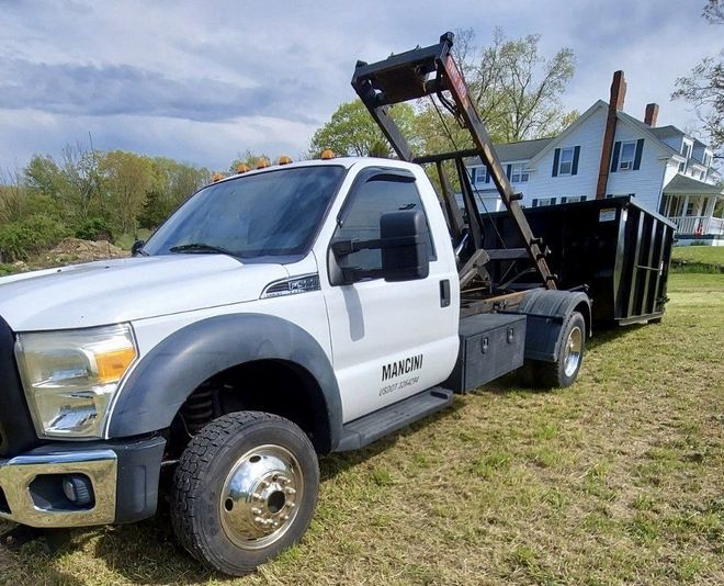 White roll-off truck with a black container on a grassy lawn, house in the background.