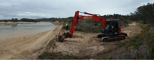 A red excavator is digging in the dirt near a body of water.