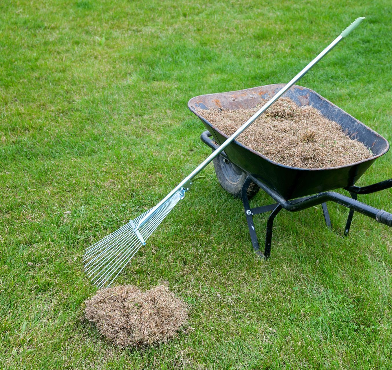 Une brouette remplie de terre et un râteau sur l'herbe.