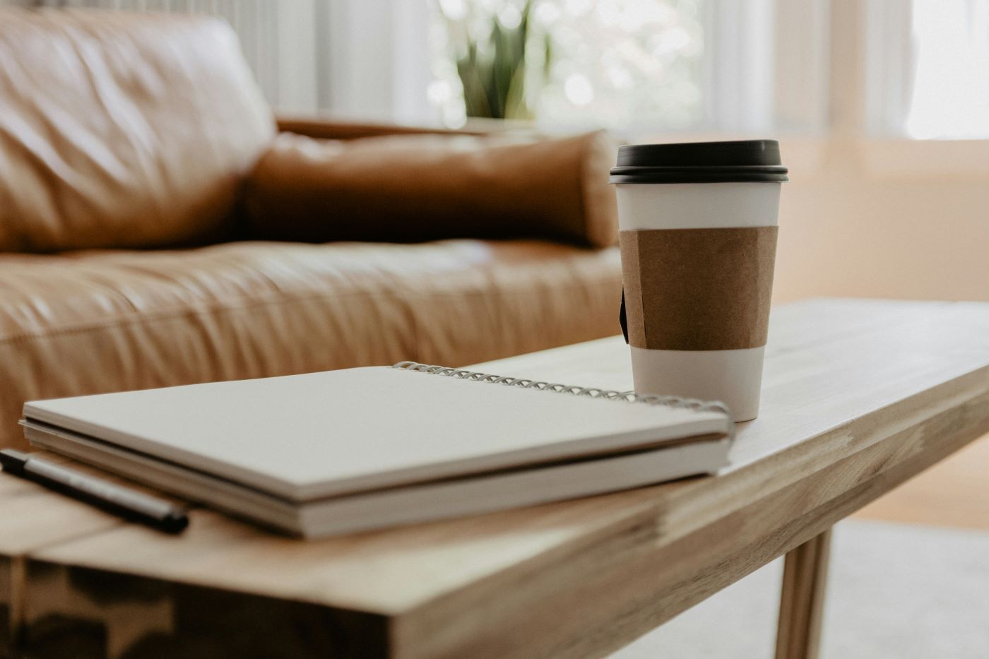 A cup of coffee is sitting on a wooden table next to a notebook and pen.