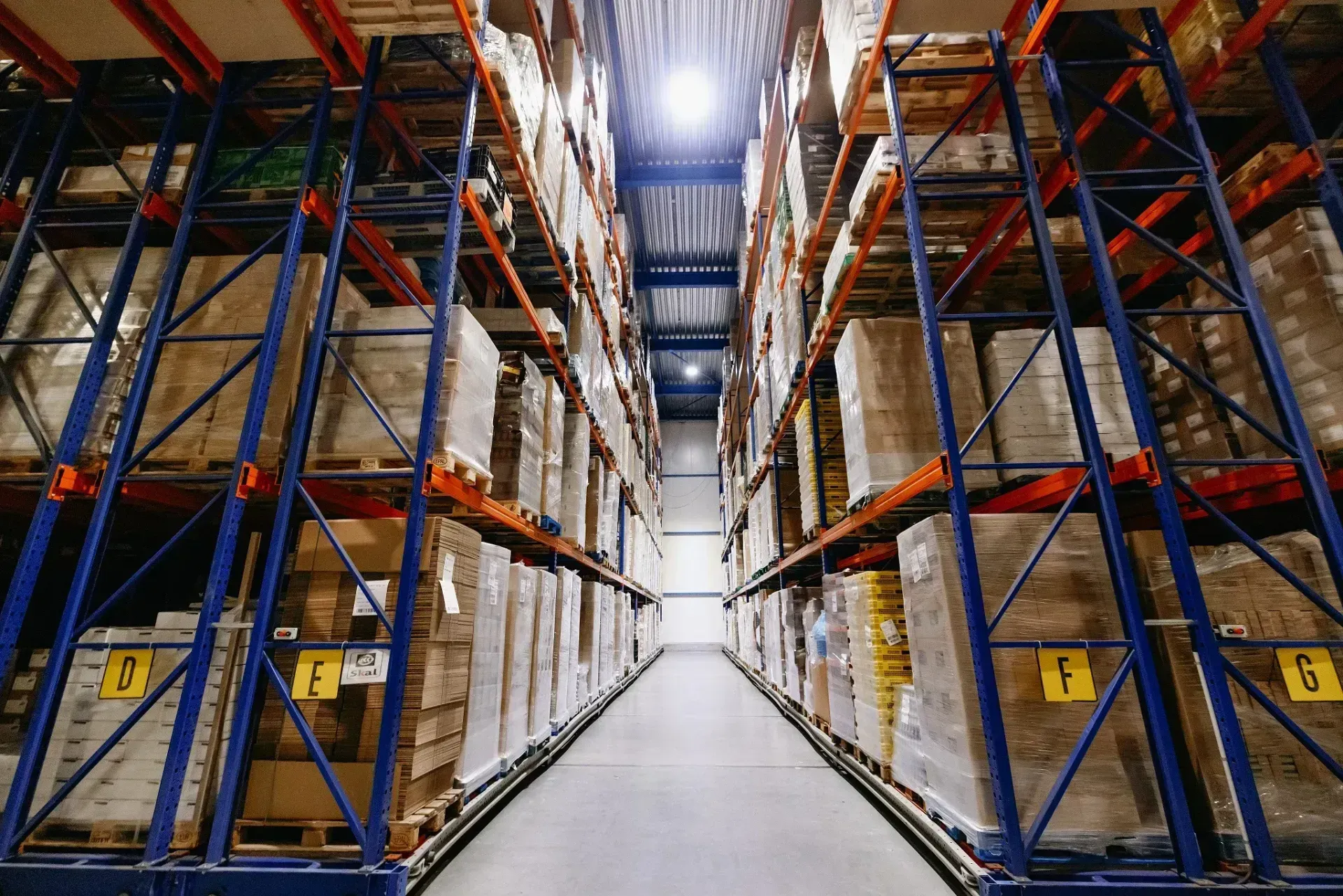 Interieur van een magazijn met rijen hoge schappen volgestapeld met pakketten; een centrale gang is zichtbaar.