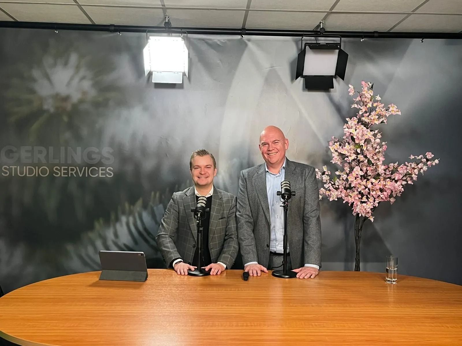 Twee mannen aan een studiotafel, achter microfoons. Grijze achtergrond met bloemenmotief, podiumverlichting.