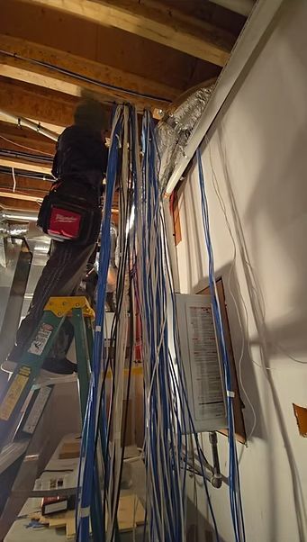 A person on a ladder is working on a bundle of wires near a wall in a basement setting.