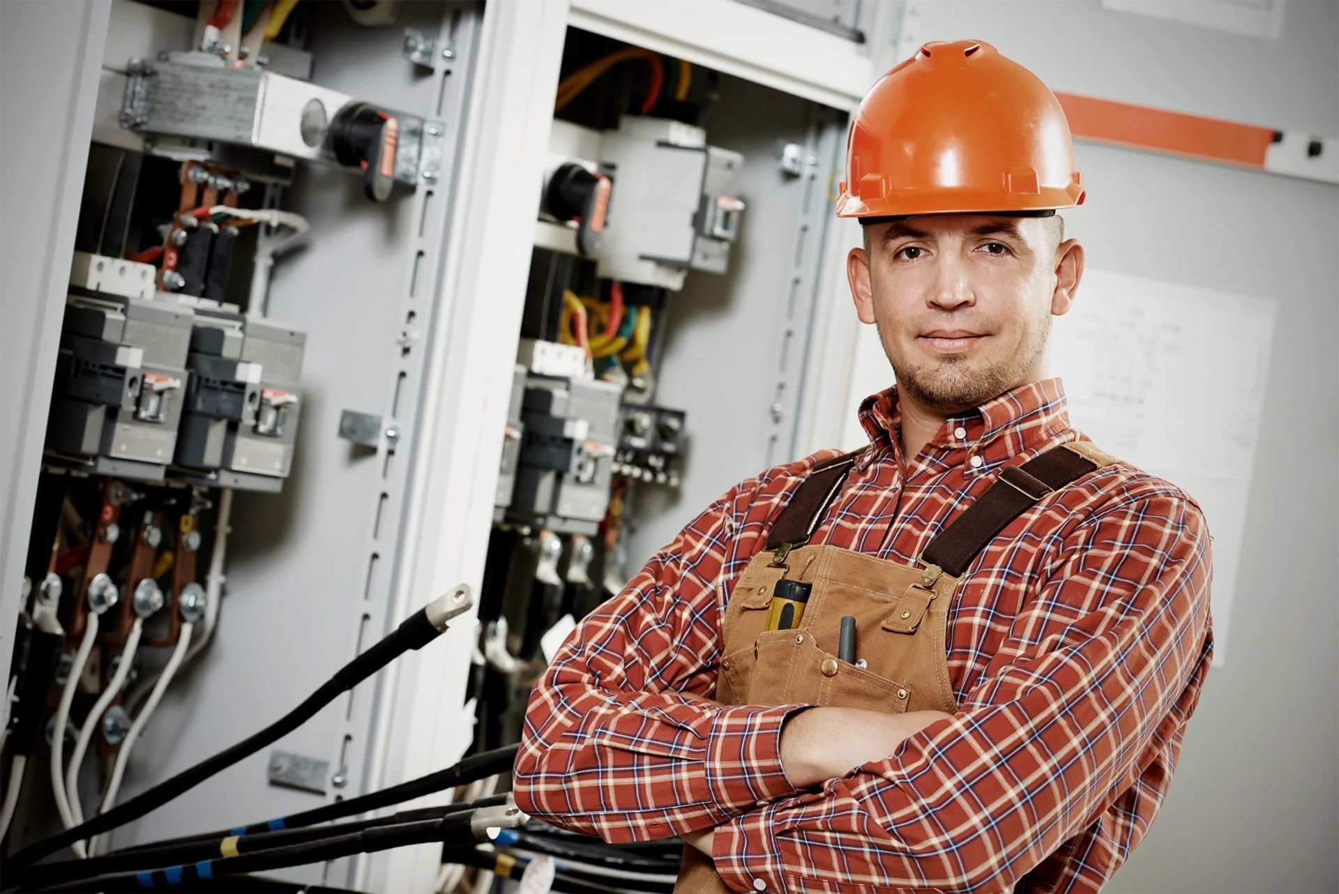 Electrician in an orange hard hat stands in front of an electrical panel, arms crossed, smiling.