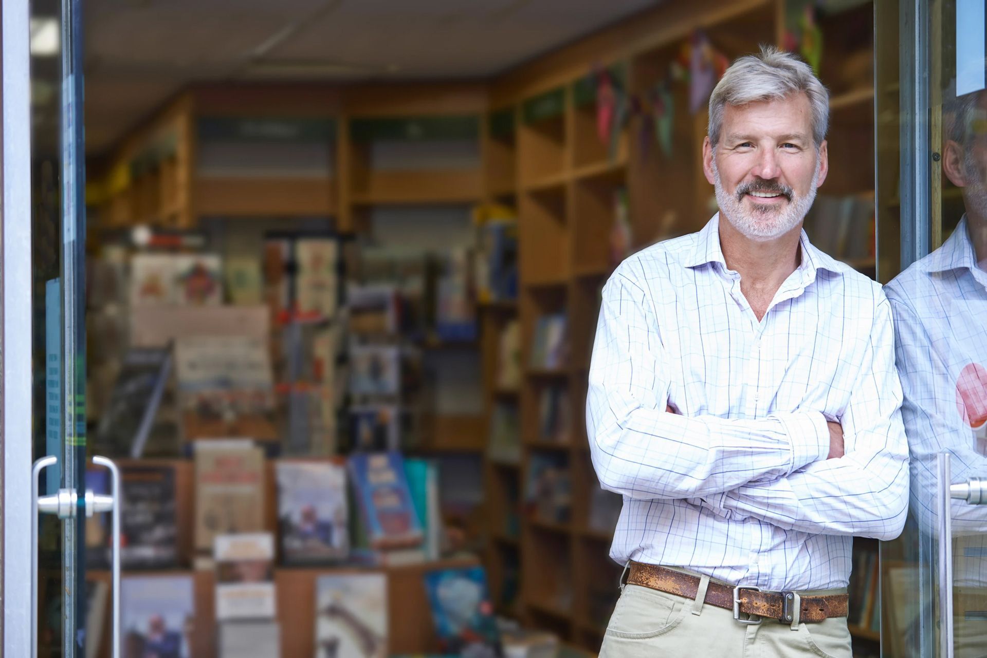 Man with gray hair smiles, arms crossed, standing in doorway of a bookstore.