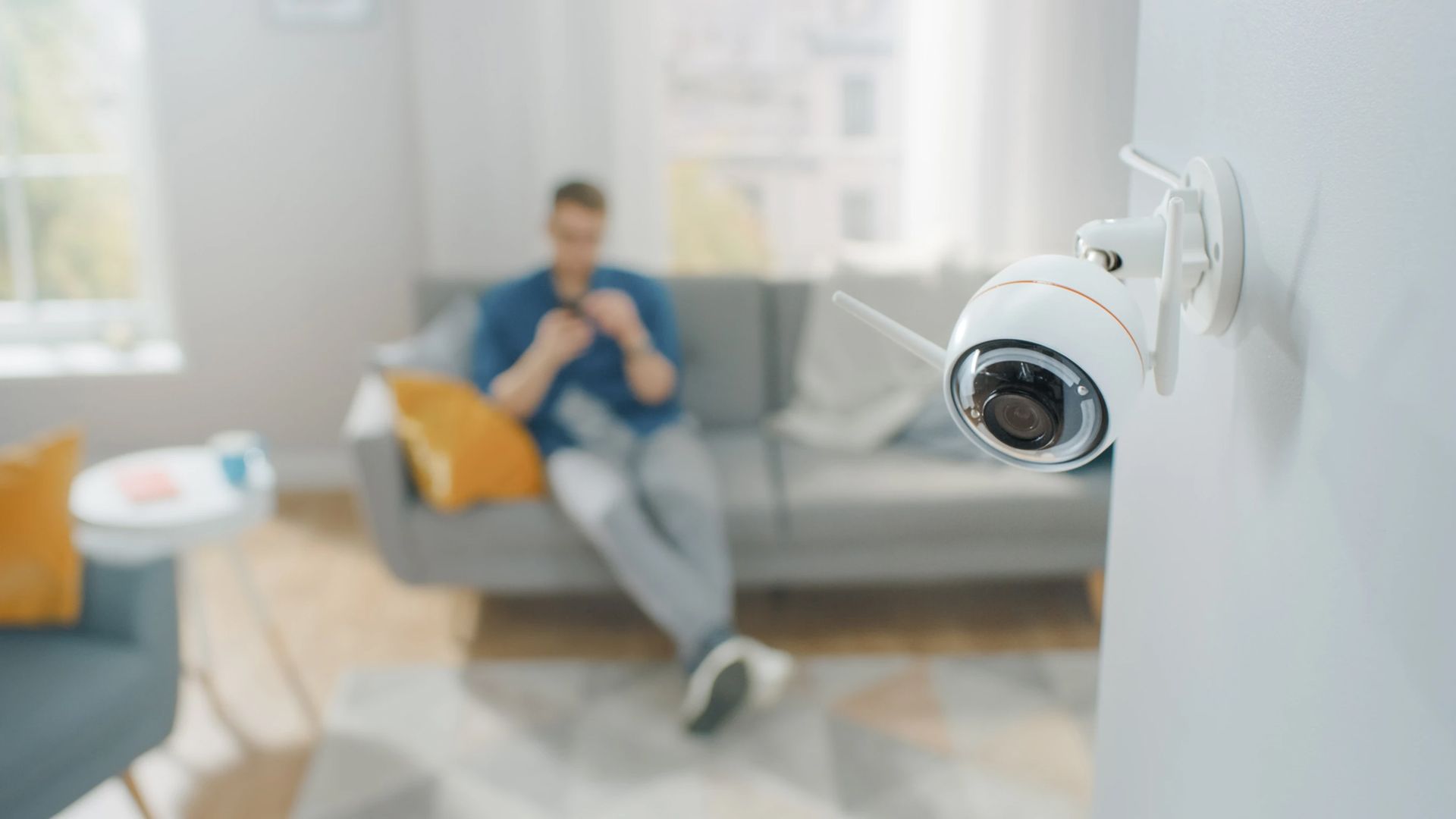 Security camera mounted on wall, overlooking a living room where a man sits on a couch, using his phone.