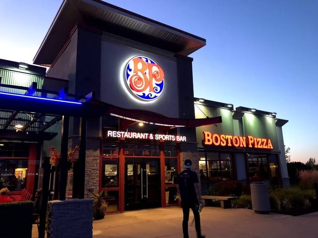 Boston Pizza restaurant exterior at dusk; illuminated sign, red and blue logo, person stands in front.