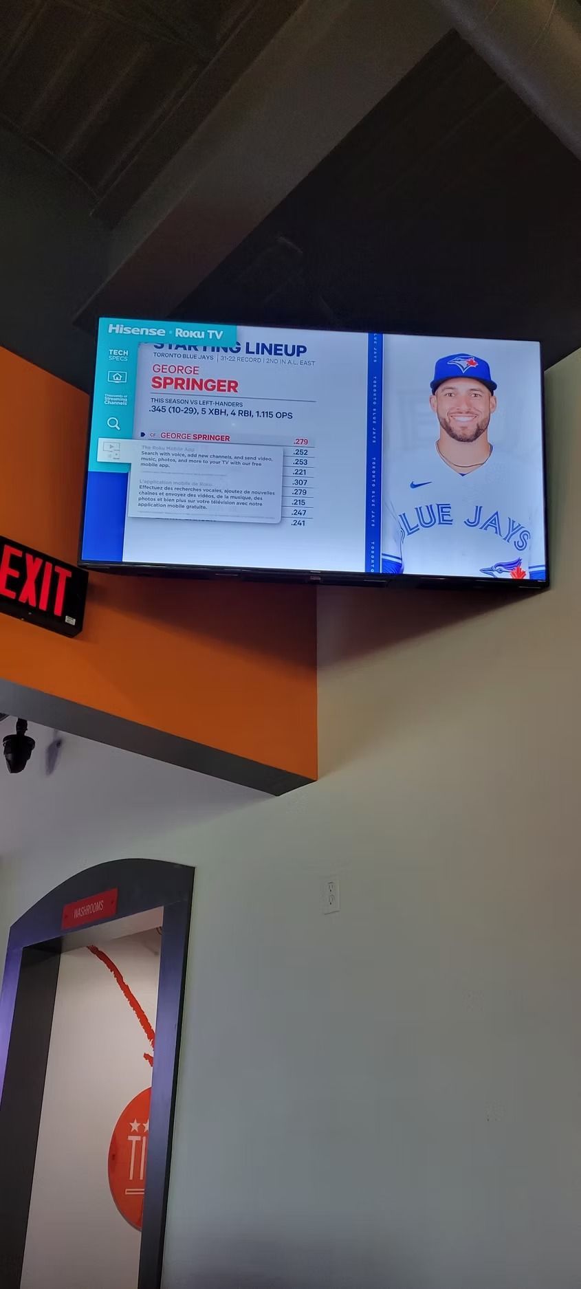 Television displaying a baseball player. A wall with an exit sign, a door, and orange and gray paint.