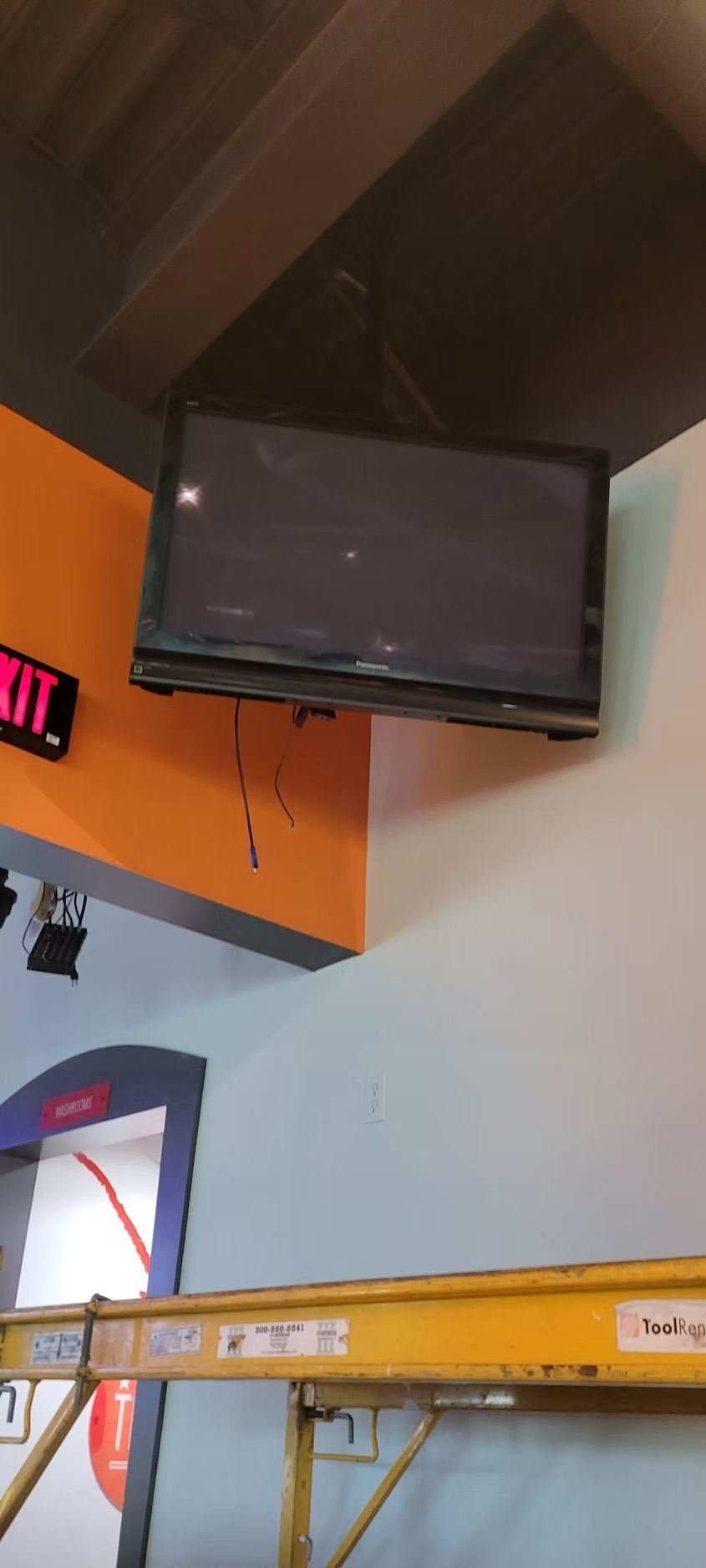 A TV mounted on a corner wall with orange and white paint. A yellow scaffold is in the foreground.