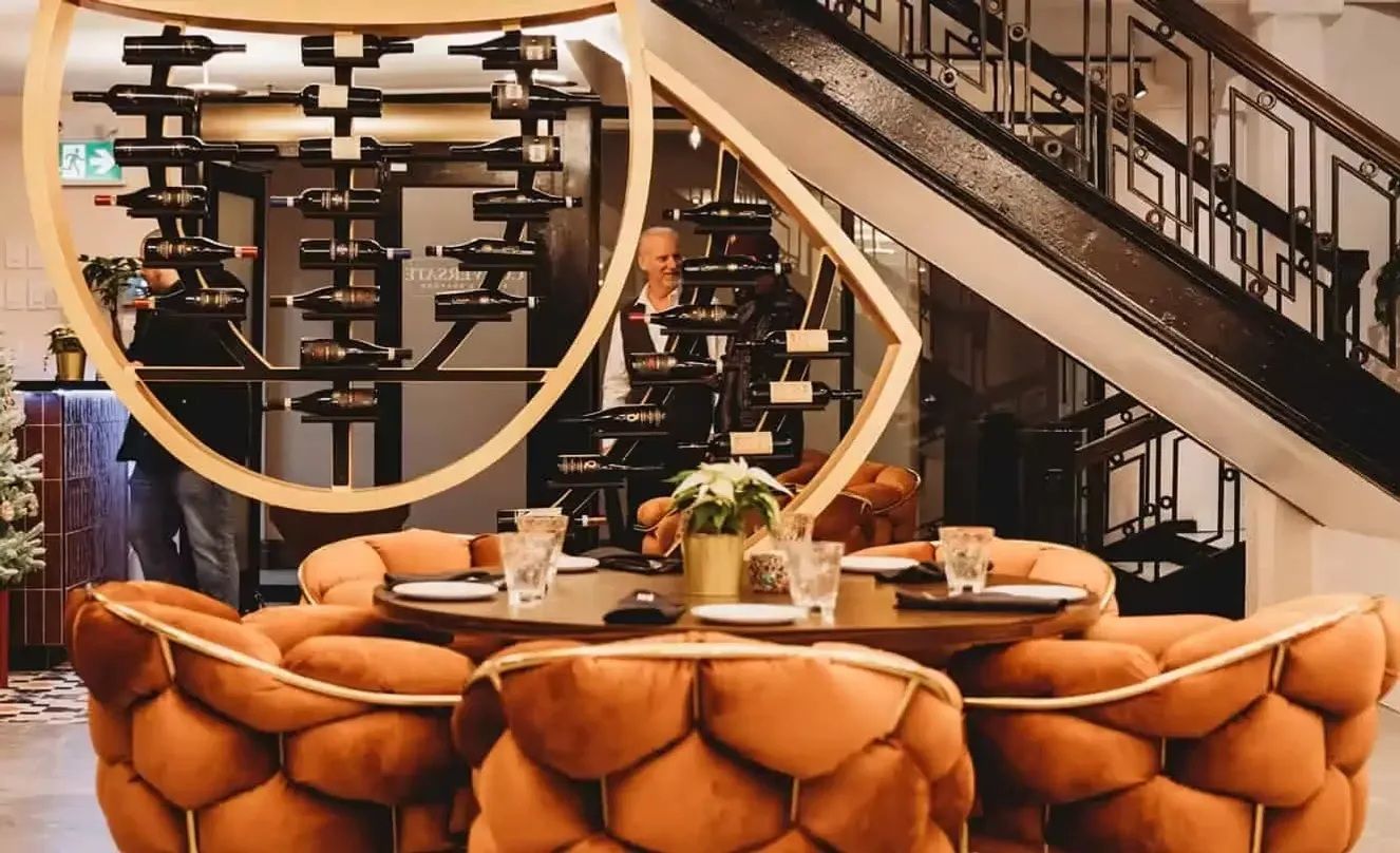 Restaurant dining area with orange chairs, round table set for guests, wine rack, and stairs.
