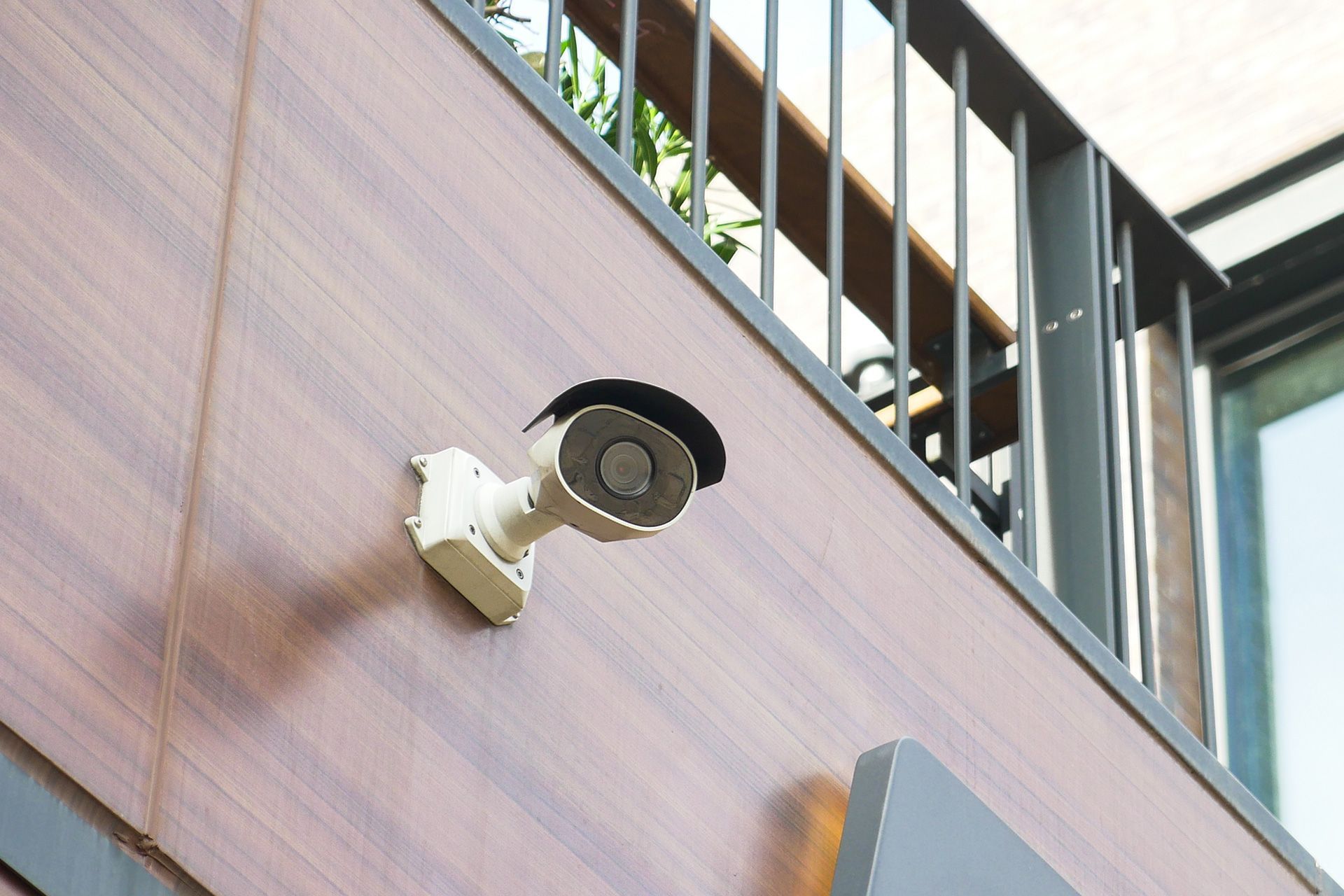 Security camera mounted on a building's exterior, angled upward. The building has brown wood paneling and a railing.