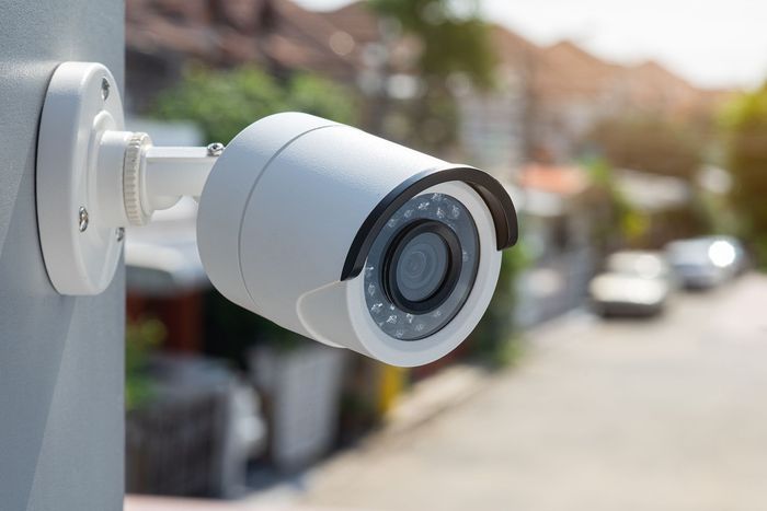 White security camera mounted on a wall, overlooking a street with cars.