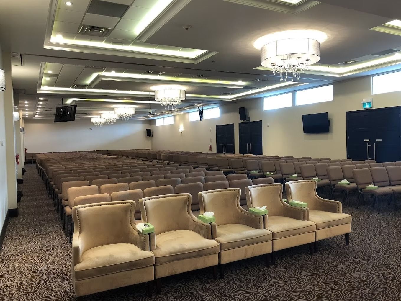 An empty auditorium with beige chairs in rows, cream accent chairs at the front, and chandeliers.