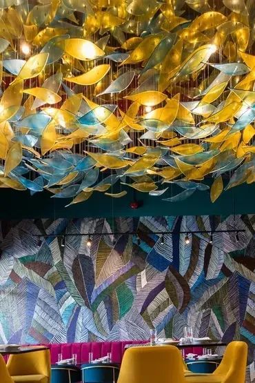 Restaurant interior with a colorful leaf-themed ceiling light fixture and wall mural. Yellow chairs and pink booth seating.