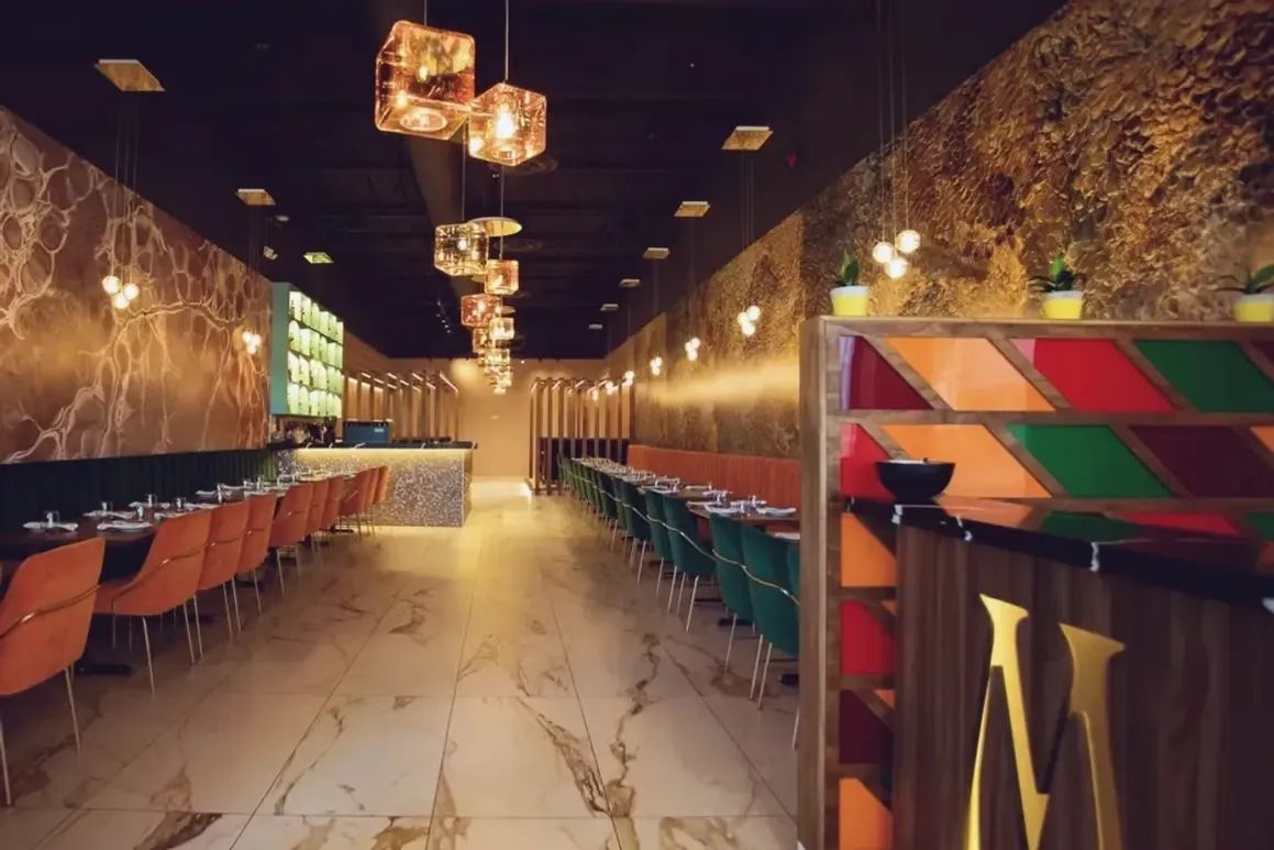 Restaurant interior with marble-patterned walls, orange and green chairs, and cube-shaped hanging lights.