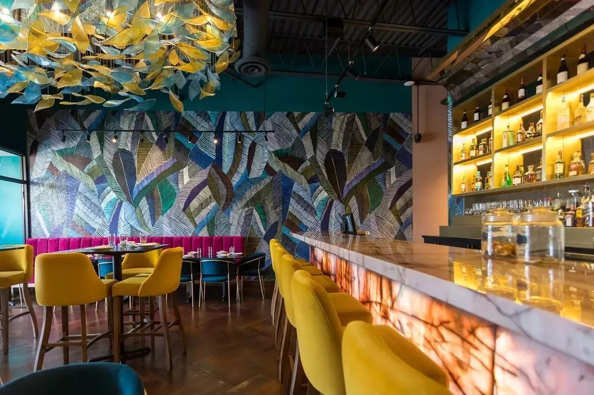 Restaurant interior with yellow chairs, bar, colorful wall mural, and shelves of liquor bottles.