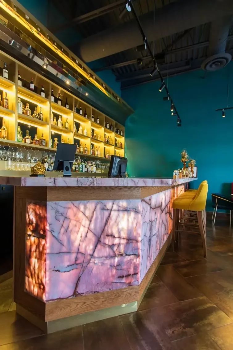 Bar with backlit pink stone front, wooden trim, bottles on shelves, and turquoise wall.