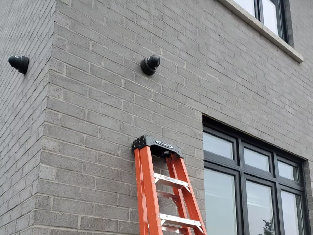 Gray brick building with a ladder leaning against it and two black pipes.