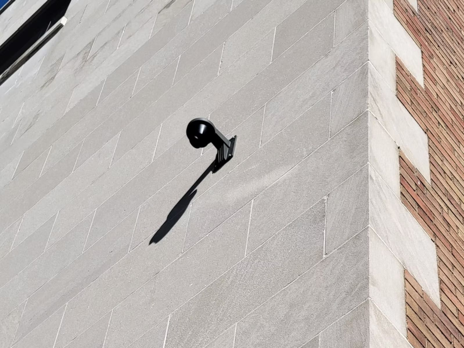 Black security light on a light gray stone wall with a brick corner; shadow cast diagonally.