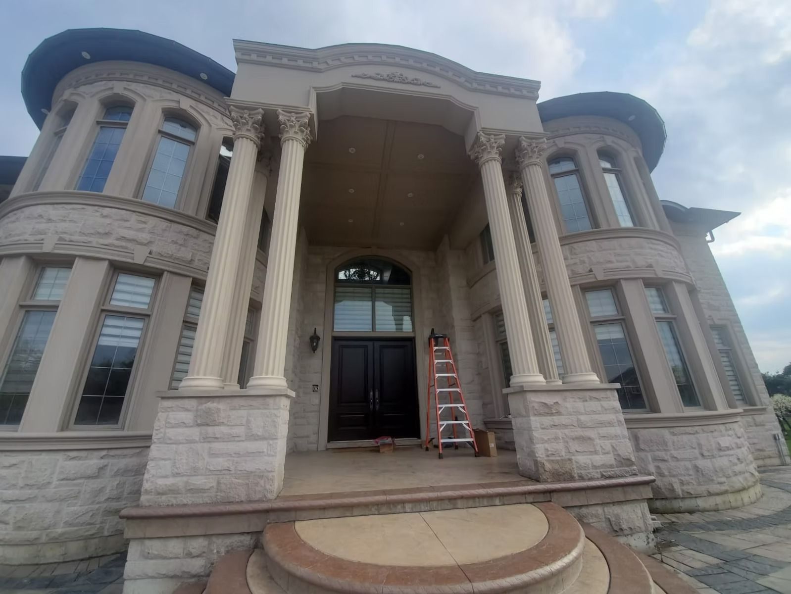 Luxurious two-story house with stone facade, columns, and curved windows. A ladder leans against the front door.