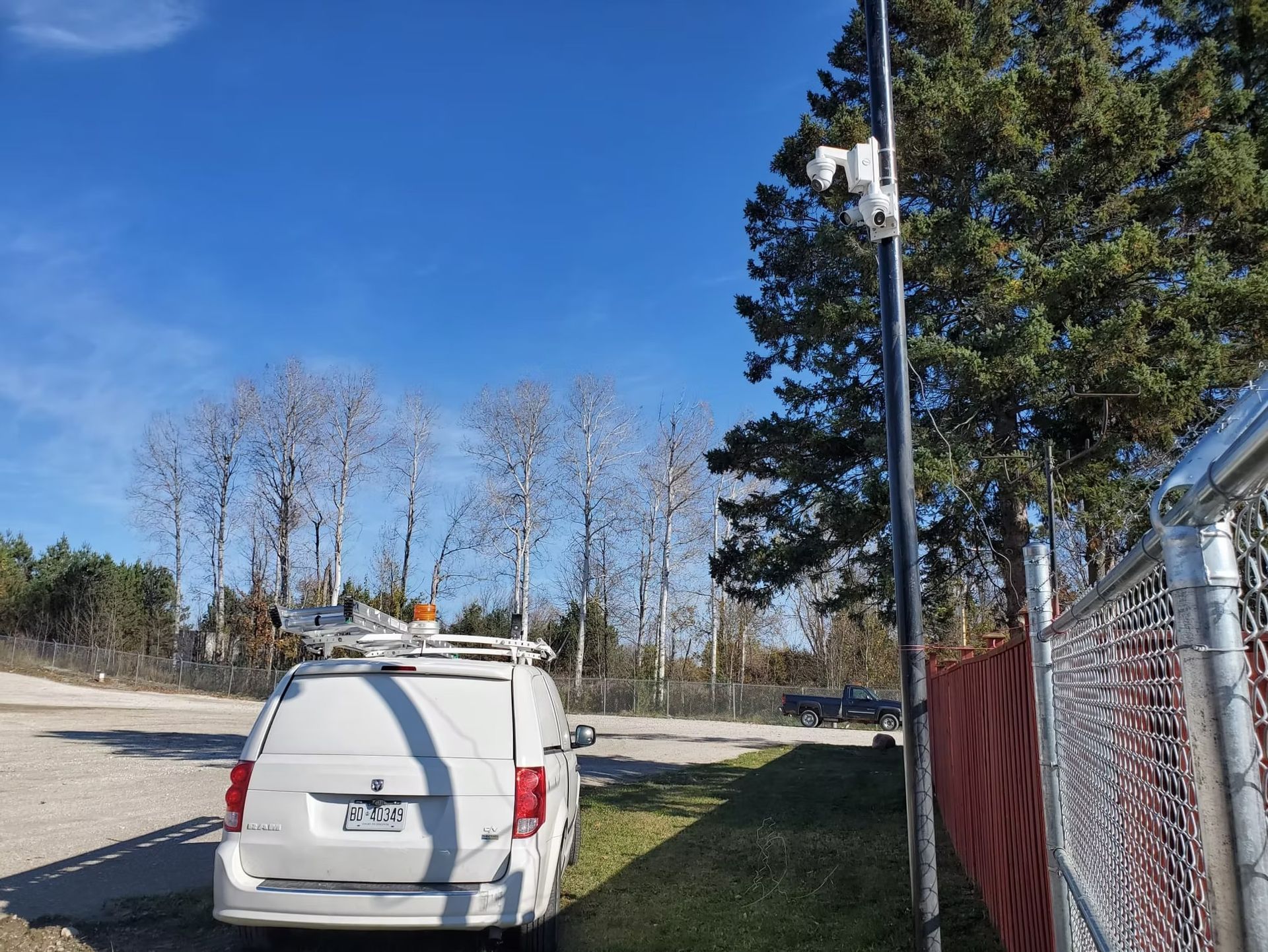 White van parked next to a black pole with equipment, fence, trees, and blue sky.