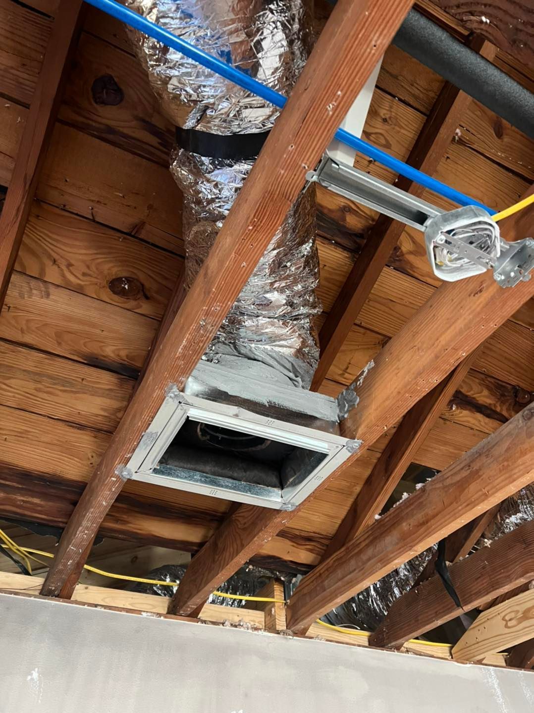 HVAC ductwork in attic with flexible silver ducting, metal vent, wooden beams and blue pipes.