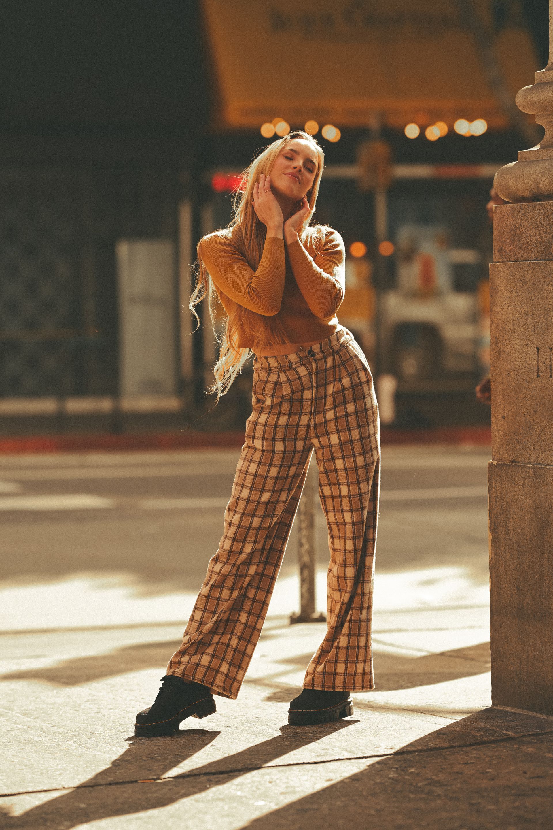 A woman in plaid pants and a yellow sweater is standing on a sidewalk.