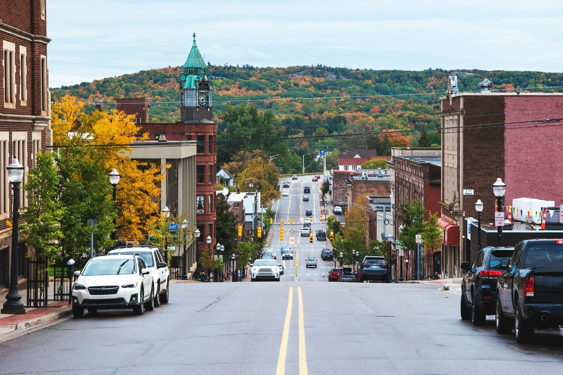 Street view of a town, vehicles on the road, buildings on either side, autumn trees, and a hill in the background.
