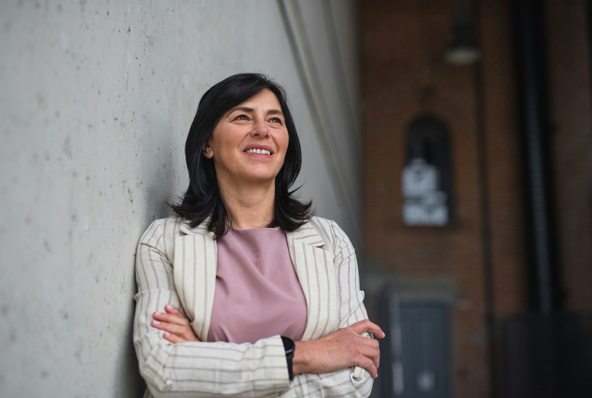 Woman in a pink top and striped blazer leans against a concrete wall, smiling and looking up.