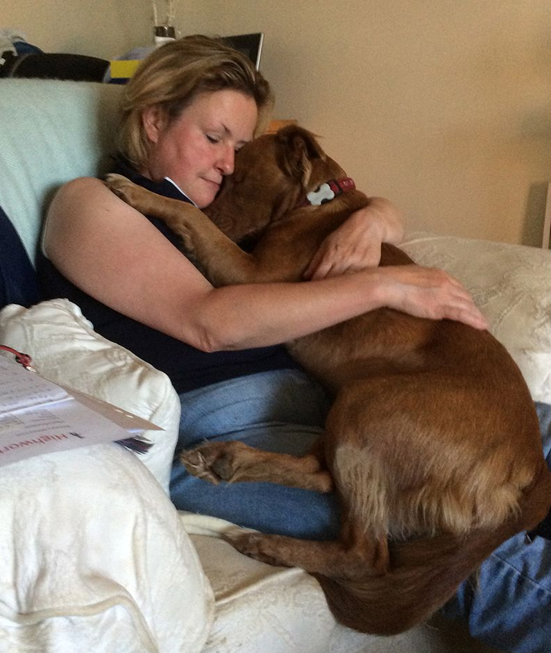 Woman on a couch hugging a brown dog in a cozy living room.