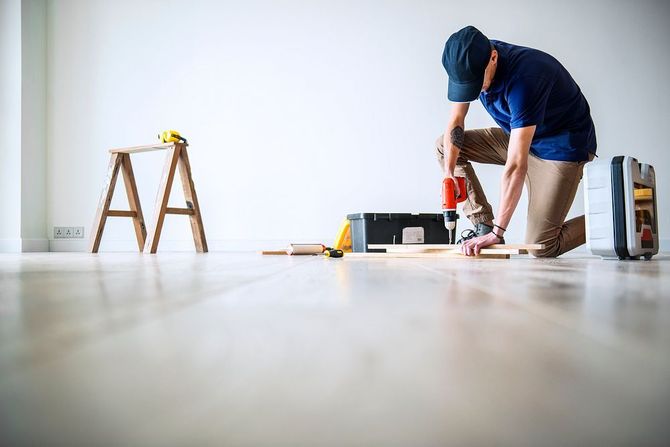 Person kneeling on floor using a power drill during home improvement, with ladder and tools nearby