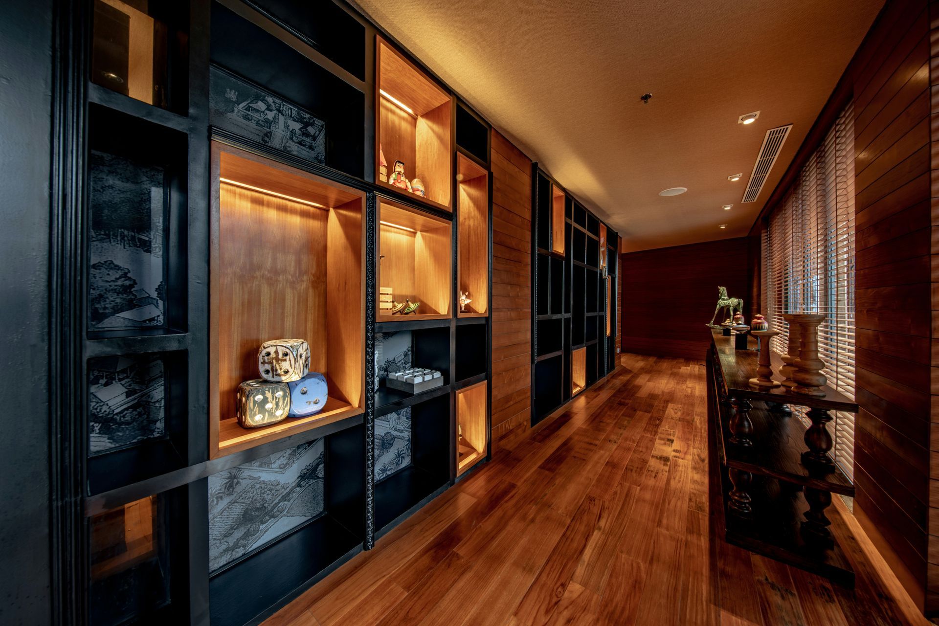 Warm-lit modern hallway with wooden floors, black shelving, and decorative wall shelves on the left.