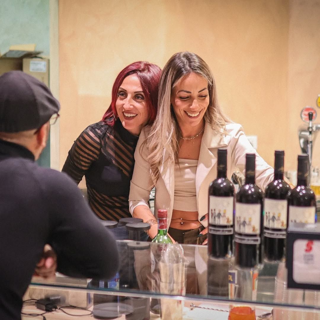 Due donne sorridono dietro il bancone di un bar, con delle bottiglie di vino davanti. Una persona è di fronte a loro.